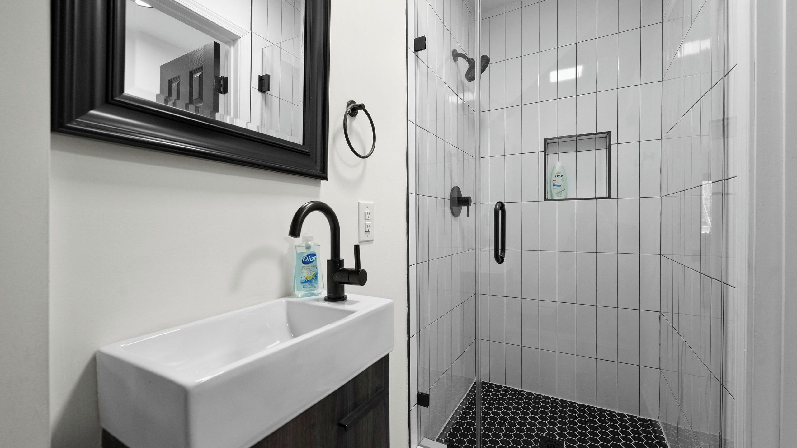 A bathroom with a white sink, black faucet, and a mirror. There is a shower area with white tiles and a small window.