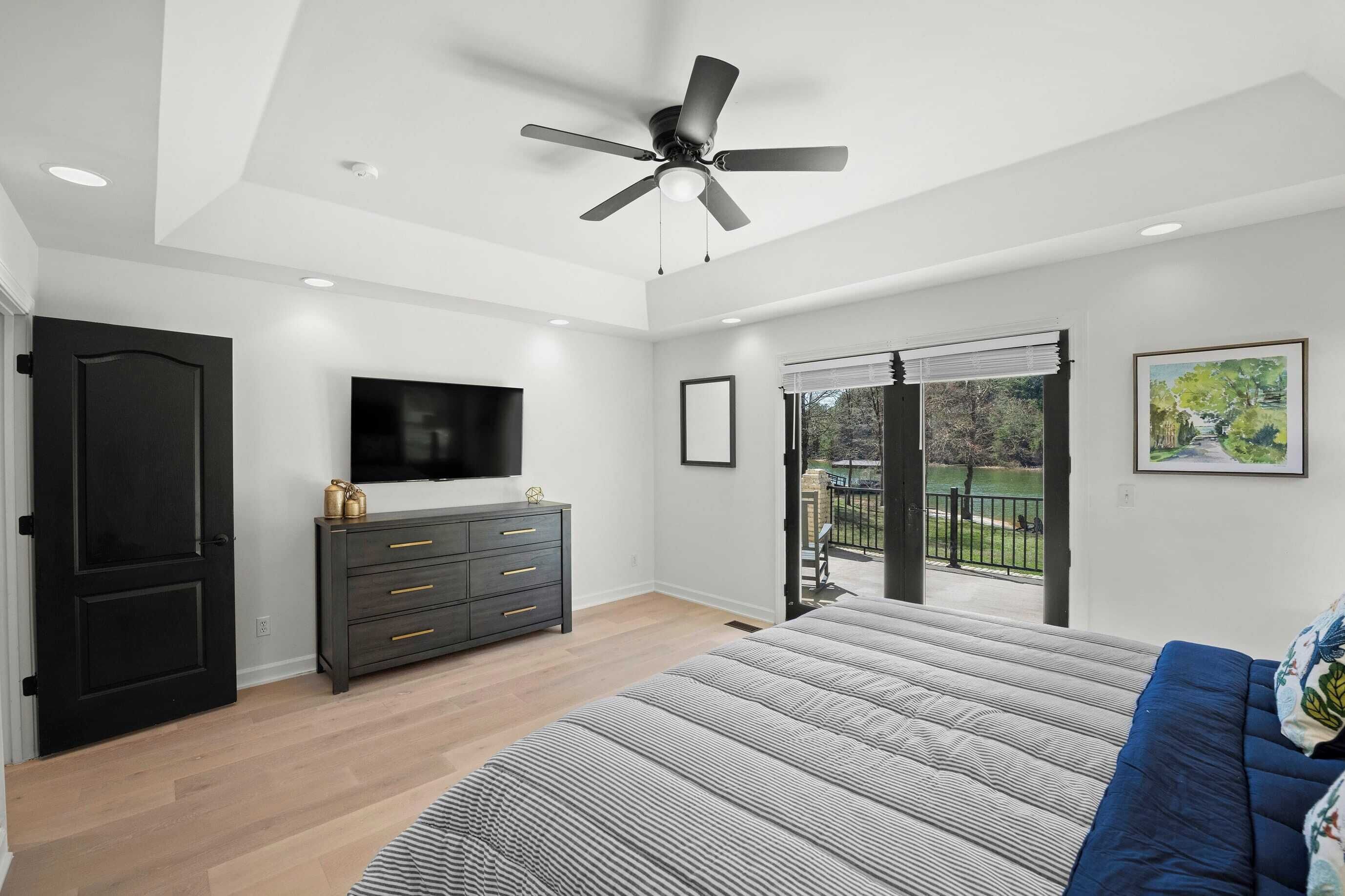 Un dormitorio con cama, ventilador de techo, televisor, cómoda y balcón con vistas.