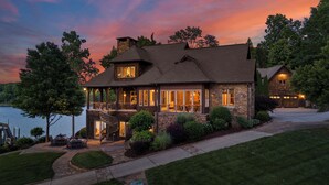 A two-story house with a stone facade, large windows, and a deck overlooking a body of water. - Sunset Shores Resort: The Grand House by Avantstay Lakefront, Spa, Private Dock+beach, Game Room (Mooresville)