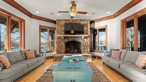 A living room with a stone fireplace, two sofas, a coffee table, and a TV mounted above the fireplace. - Sunset Shores Resort: The Grand House by Avantstay Lakefront, Spa, Private Dock+beach, Game Room (Mooresville)