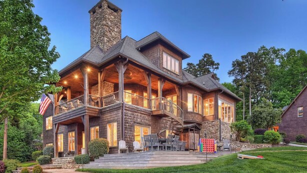 A large, multi-story house with a stone chimney, a covered porch with outdoor seating, and a flag. - Sunset Shores Resort: The Grand House by Avantstay Lakefront, Spa, Private Dock+beach, Game Room (Mooresville)