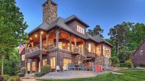 A large, multi-story house with a stone chimney, a covered porch with outdoor seating, and a flag. - Sunset Shores Resort: The Grand House by Avantstay Lakefront, Spa, Private Dock+beach, Game Room (Mooresville)