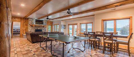Une pièce spacieuse équipée d’une table de ping-pong, d’un coin salon, d’une cheminée en pierre et de grandes fenêtres offrant une vue panoramique.