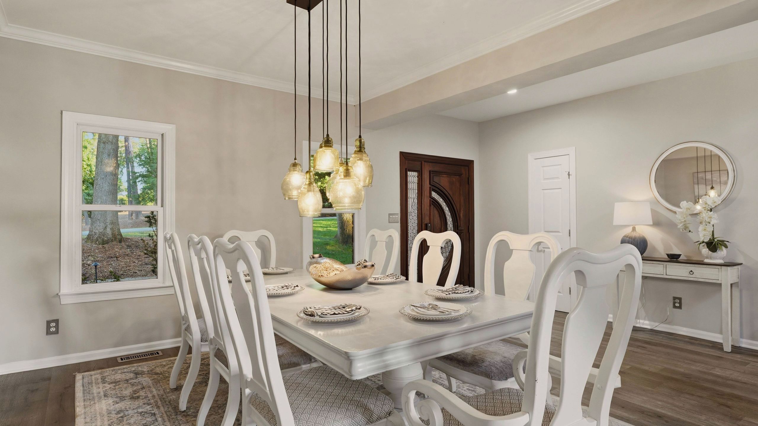A dining room with a large table, white chairs, a chandelier, and a view of trees through a window.