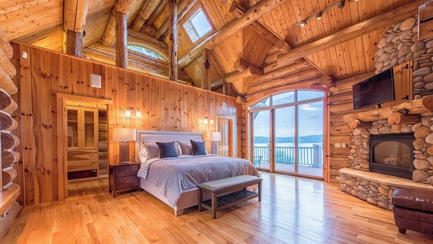 A log cabin bedroom with a large bed, a stone fireplace, and a view of the mountains. - Azalea Retreat by Avantstay Sleeps 34 & Pool (Sherrills Ford)