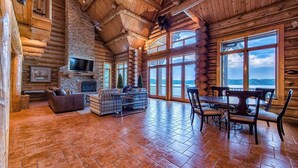 A log cabin interior with a stone fireplace, a television, and a dining area with a view of the mountains. - Azalea Retreat by Avantstay Sleeps 34 & Pool (Sherrills Ford)