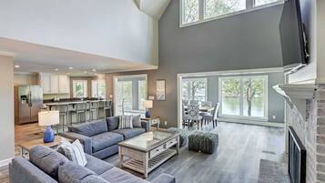 A spacious living area with a high ceiling, a kitchen in the background, and large windows offering views of the outdoors.