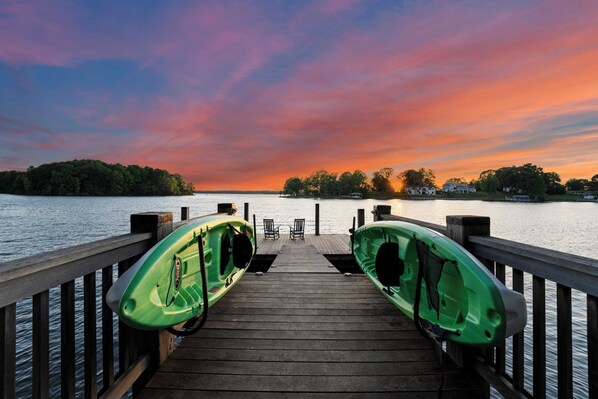 Un muelle de madera con dos kayaks verdes y una fila de sillas mirando una puesta de sol sobre el agua.