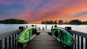 Un muelle de madera con kayaks y sillas, que se extiende hacia una masa de agua con árboles y casas al fondo.