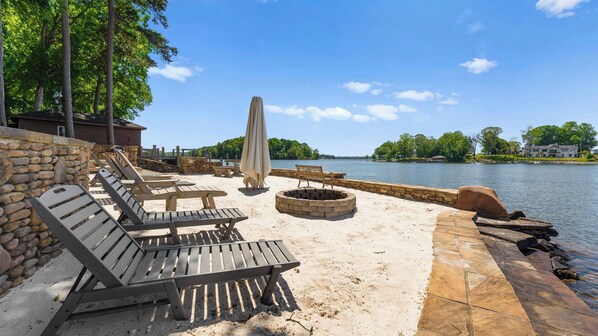 Un patio junto al lago con sillones de madera, una fogata y un muro de piedra.