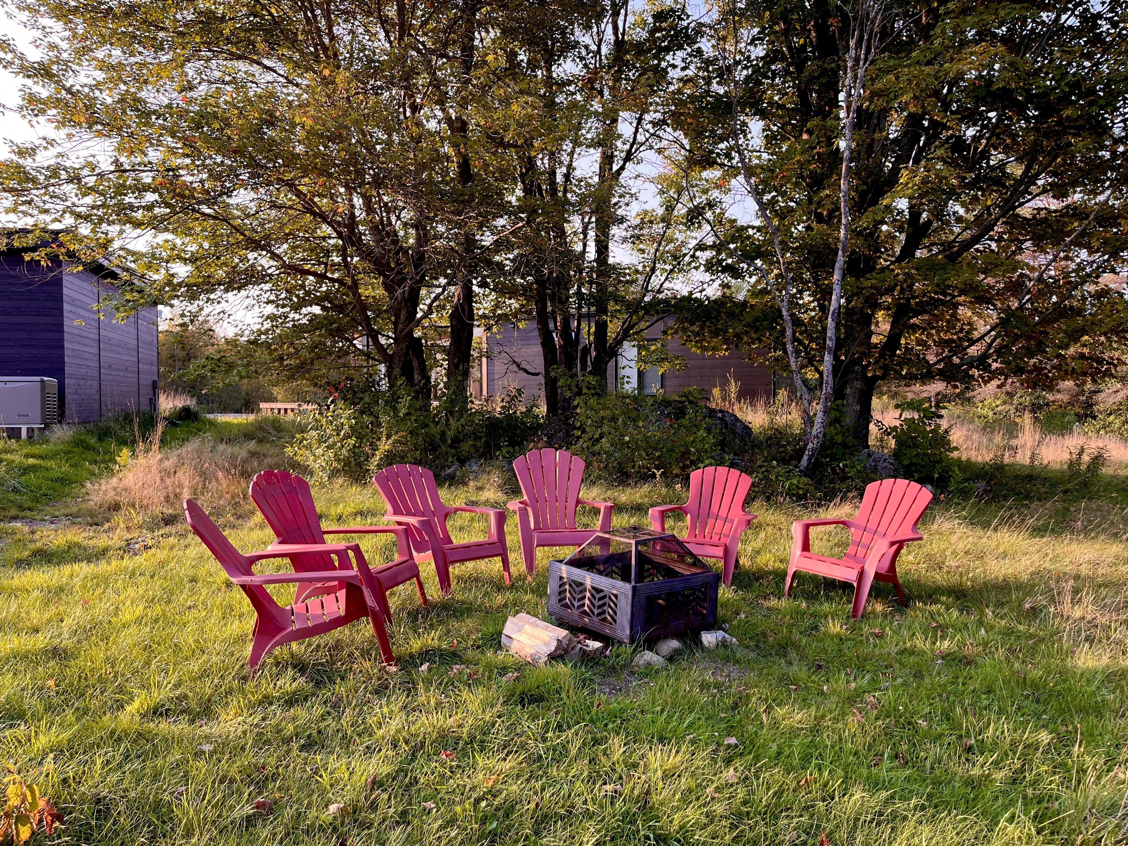 A group of red Adirondack chairs arranged around a fire pit in a grassy outdoor area with trees and a building in the background.