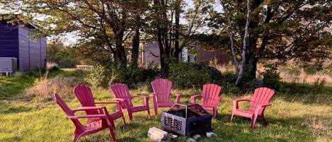 A group of red Adirondack chairs arranged around a fire pit in a grassy outdoor area with trees and a building in the background.