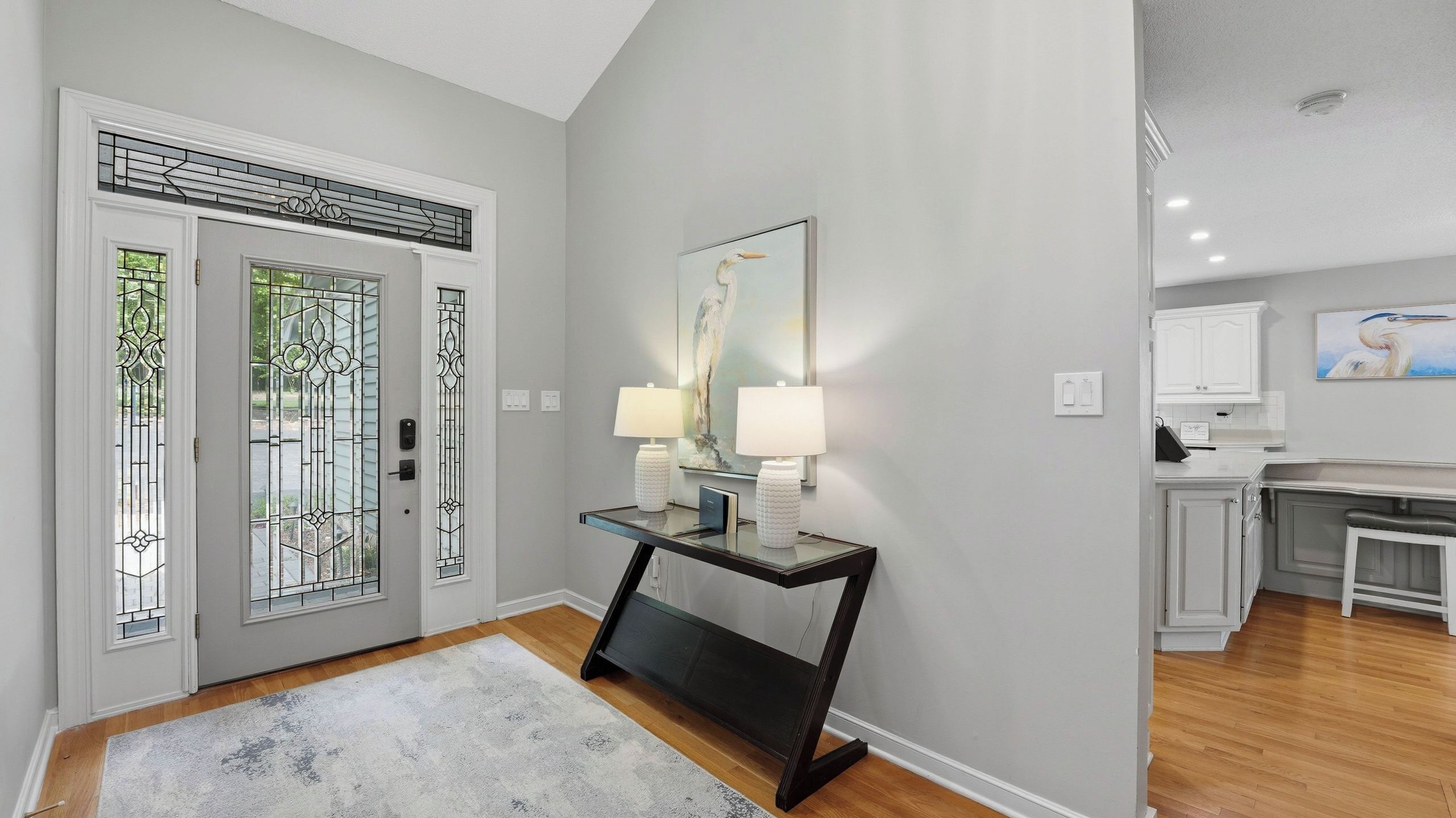 A hallway with a double door, a console table with two lamps, a framed artwork on the wall, and a kitchen area in the background.