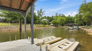 Un muelle con barcos, una zona techada y un lago con árboles y casas al fondo.