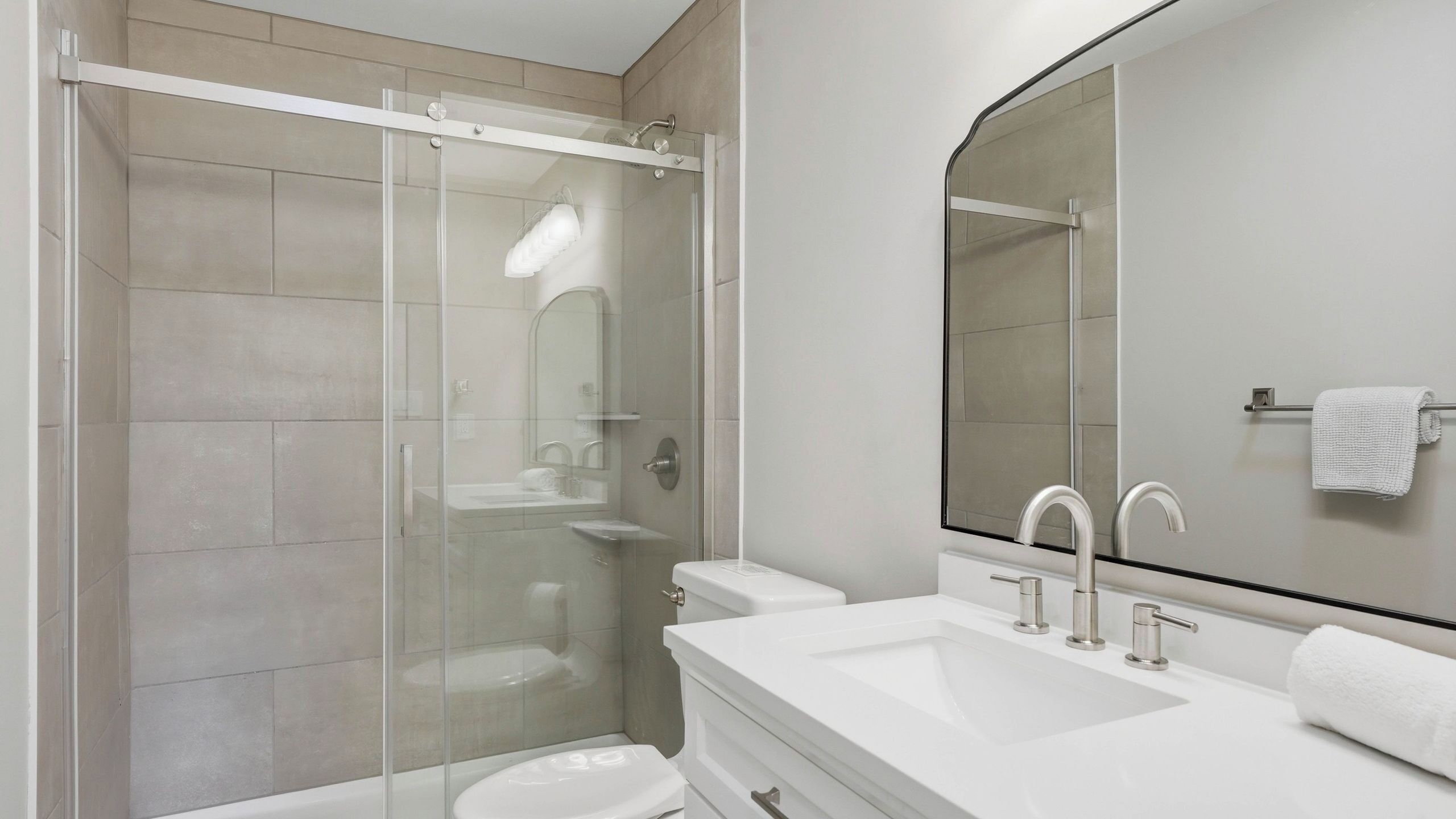 Une salle de bain moderne avec une douche à parois de verre, un lavabo blanc doté de deux robinets et un grand miroir.