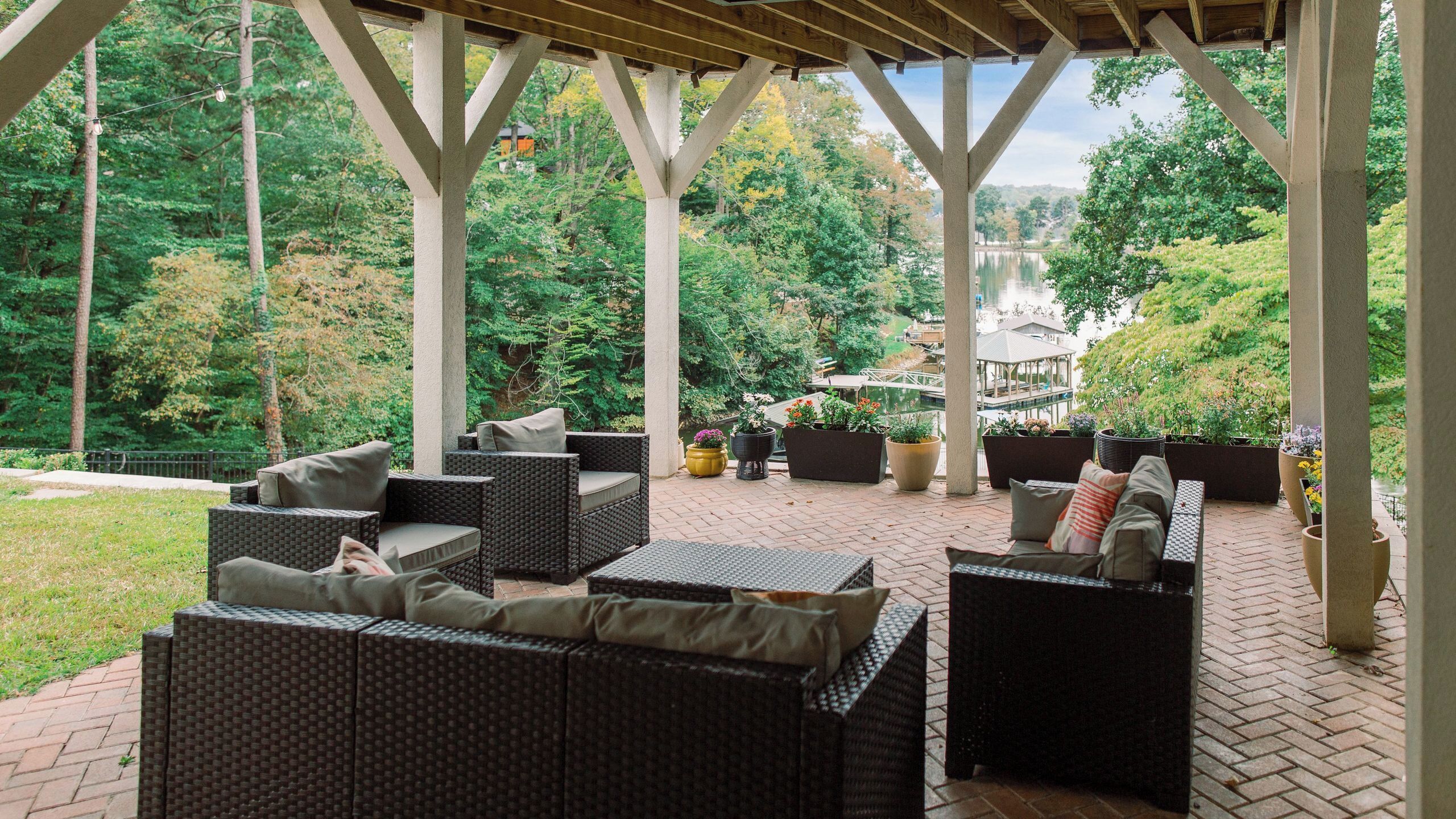 An outdoor patio with wicker furniture, potted plants, and a view of a lake and trees.
