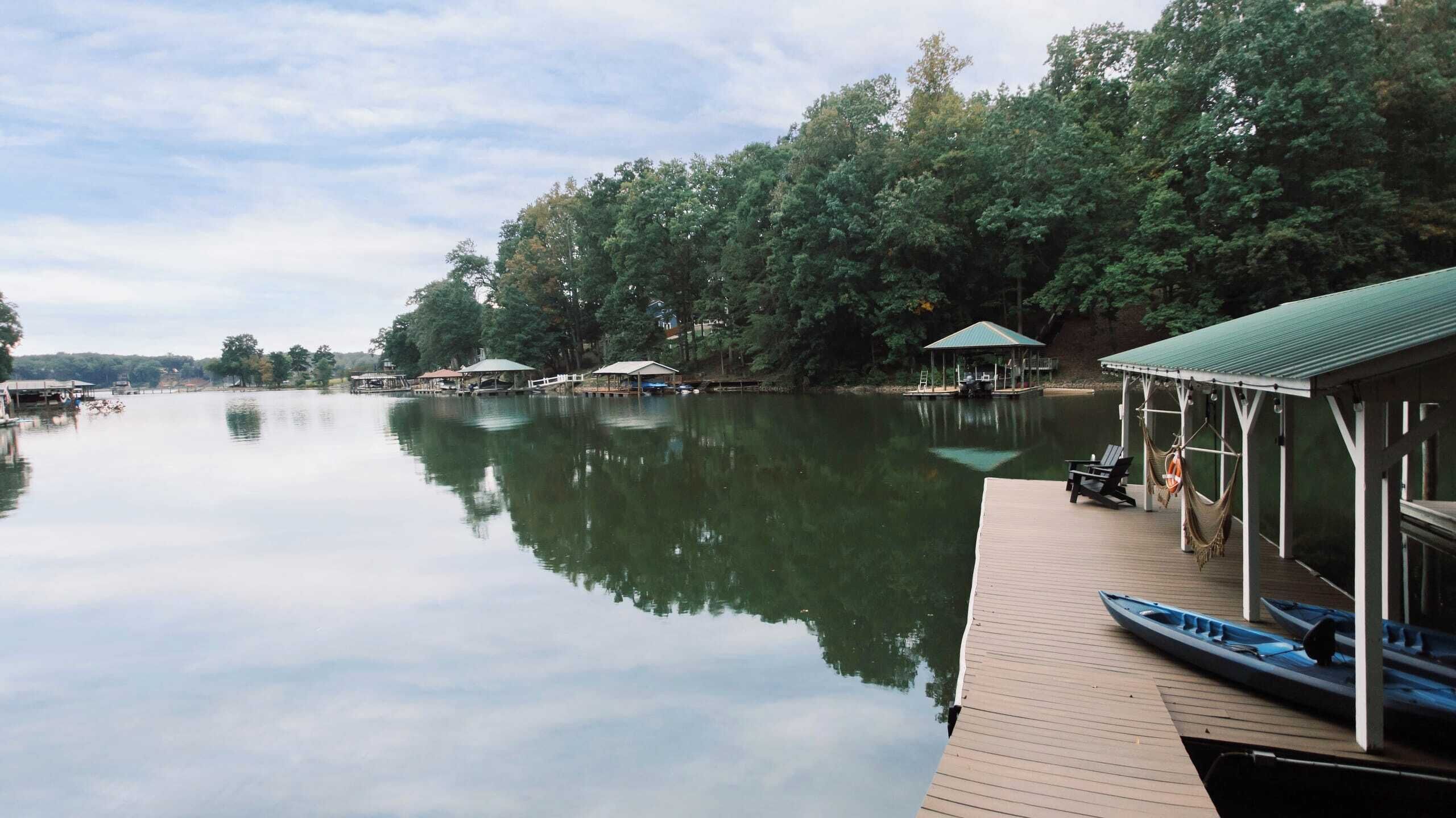 A calm lake with a wooden dock, a covered seating area, and a small boat.