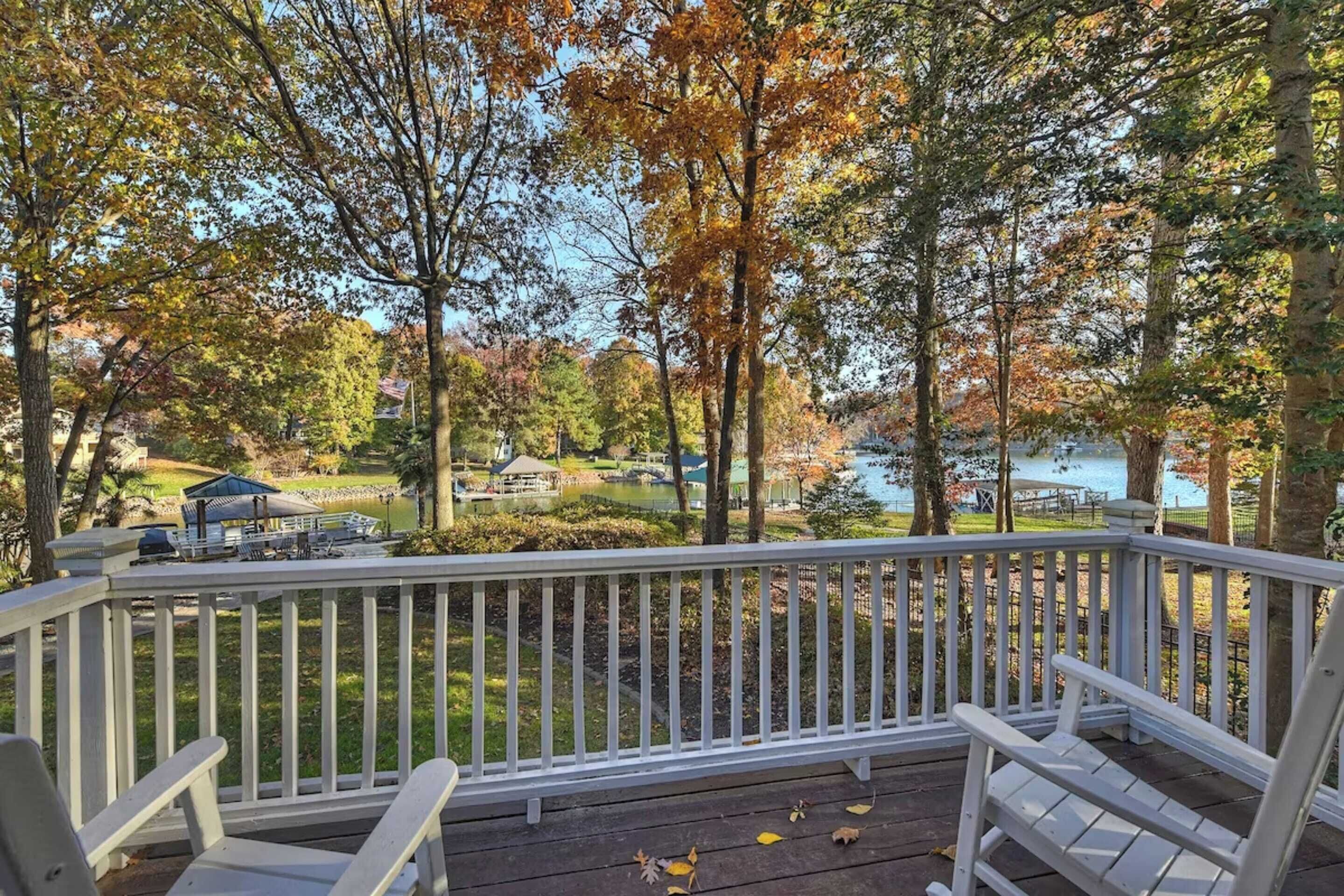 A wooden deck with white railings, two Adirondack chairs, and a view of a lake with trees in autumn colors.