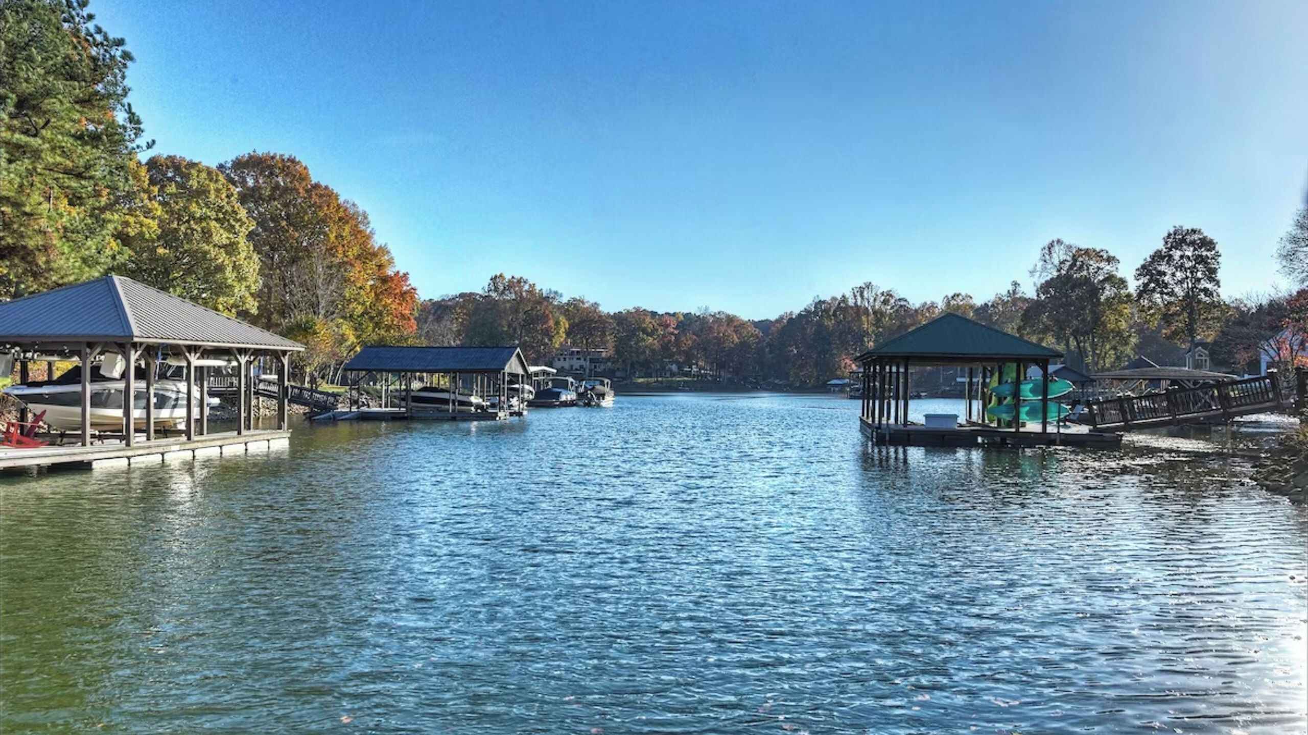 A lake with docks and covered structures, boats, and trees with autumn foliage.
