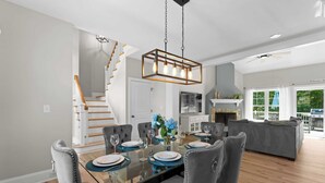 A modern dining area with a glass table, grey upholstered chairs, and a hanging light fixture. - Making Waves by Avantstay Sleeps 20 + Pool (Denver)