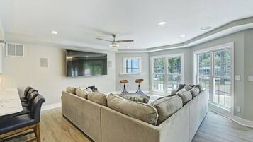 A modern living room with a sectional sofa, a flat-screen TV, a ceiling fan, and large windows.