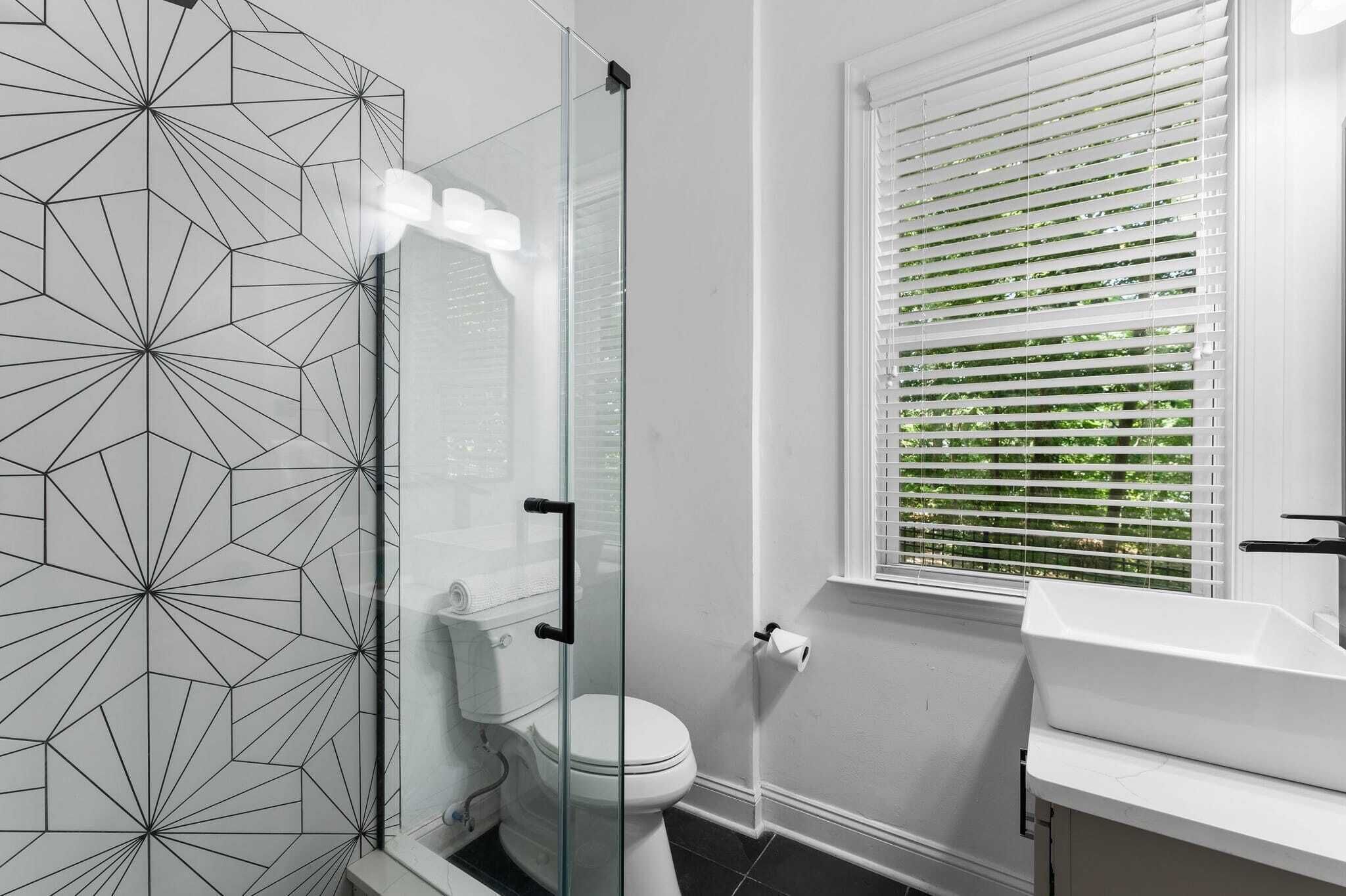 A modern bathroom with a geometric-patterned wall, a glass shower enclosure, a toilet, and a window with blinds.