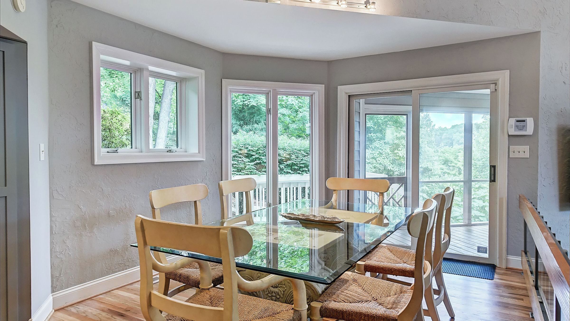 A dining area with a glass table and chairs, large windows, and a view of greenery.