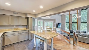A modern kitchen with a marble countertop, wooden stools, and a view of the outdoors through large windows.