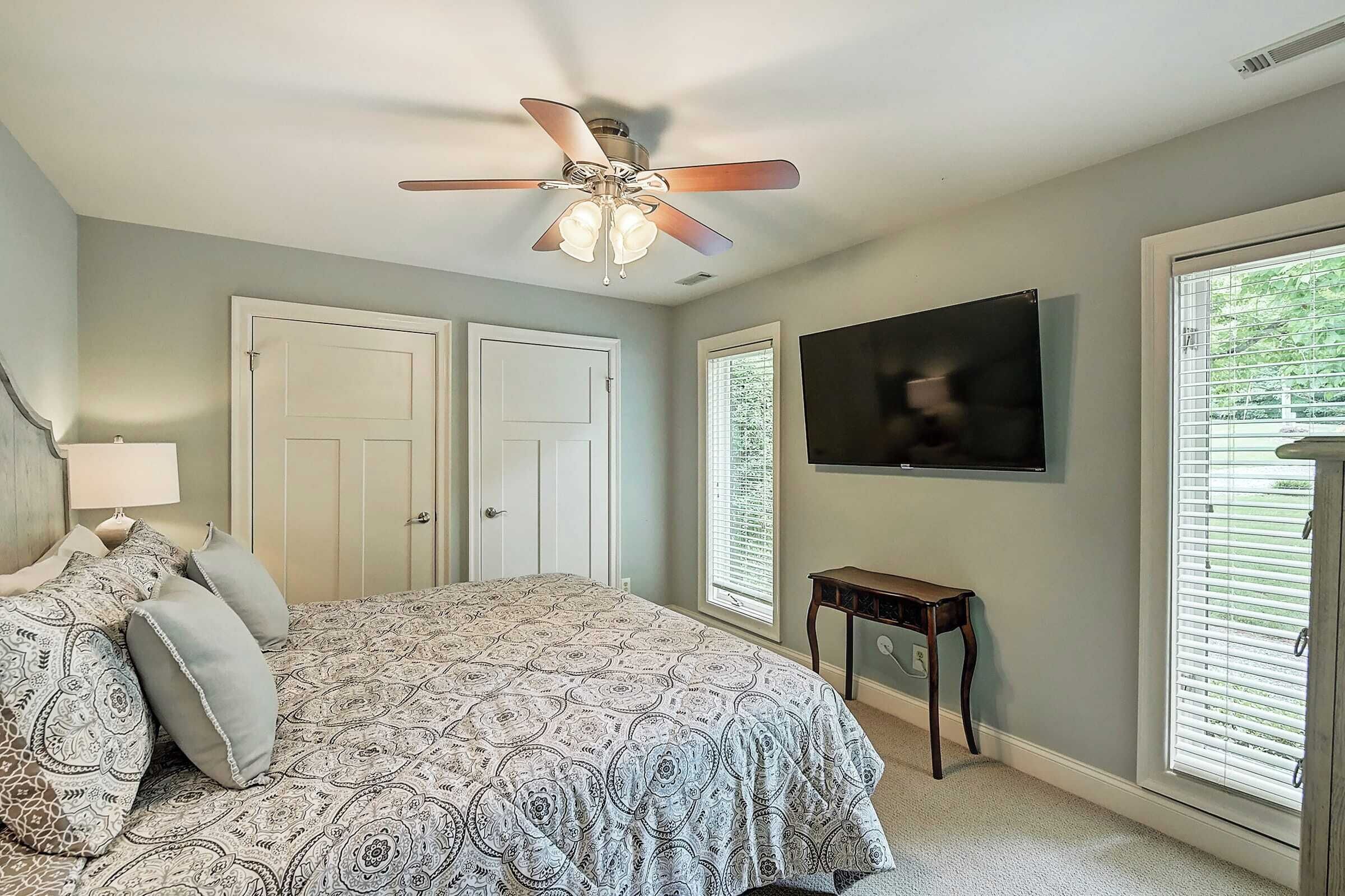 A bedroom with a bed, a ceiling fan, a TV, and a window with blinds.