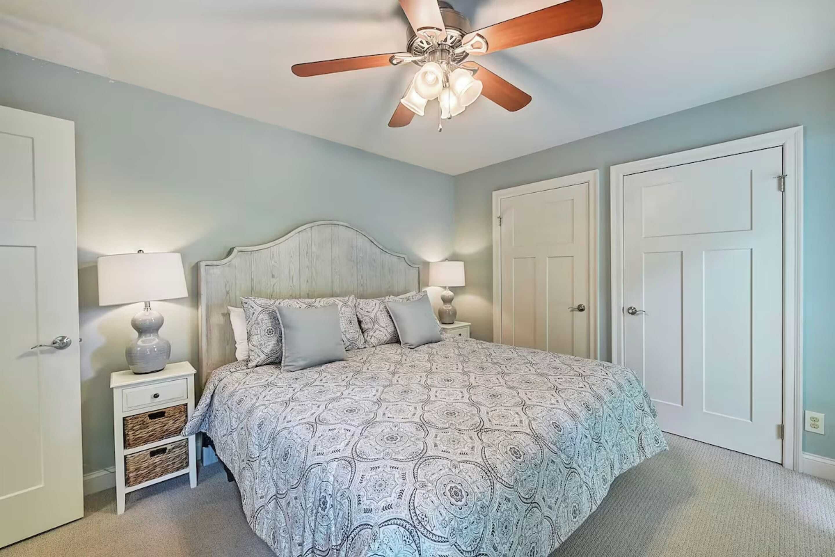 A bedroom with a bed, two bedside tables with lamps, a ceiling fan, and two doors.