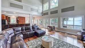 A spacious living room with a large leather sectional sofa, a dining area with a table and chairs, and large windows offering a view of the outdoors.