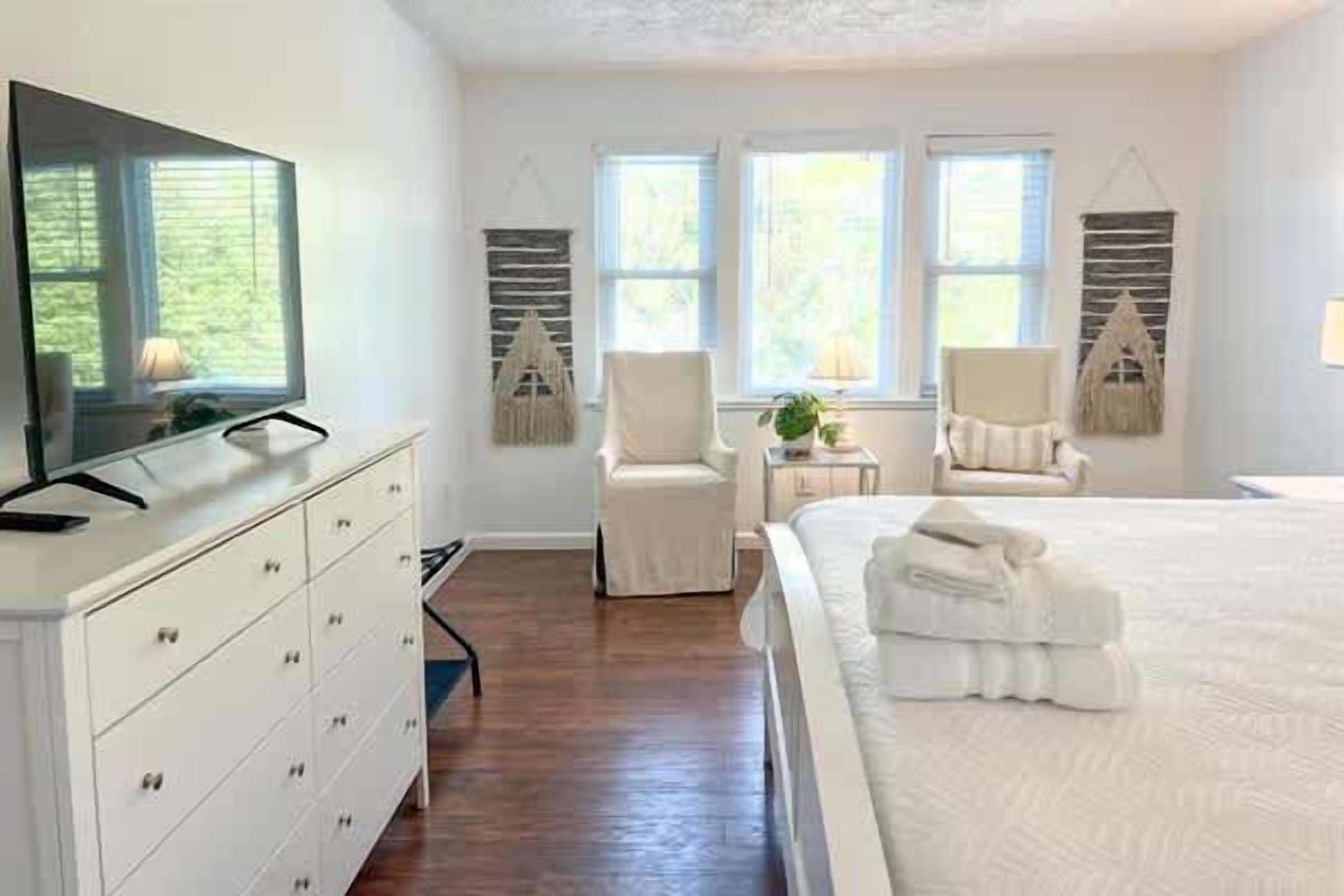 A bedroom with a bed, a TV on a dresser, two chairs, and a small table with a lamp.