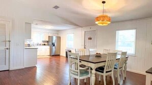 A dining area with a wooden table and white chairs, a hanging light fixture, and a view of the kitchen in the background. - Golf Cart Private pool Bay and Beach Access Pet Friendly (St. George Island)