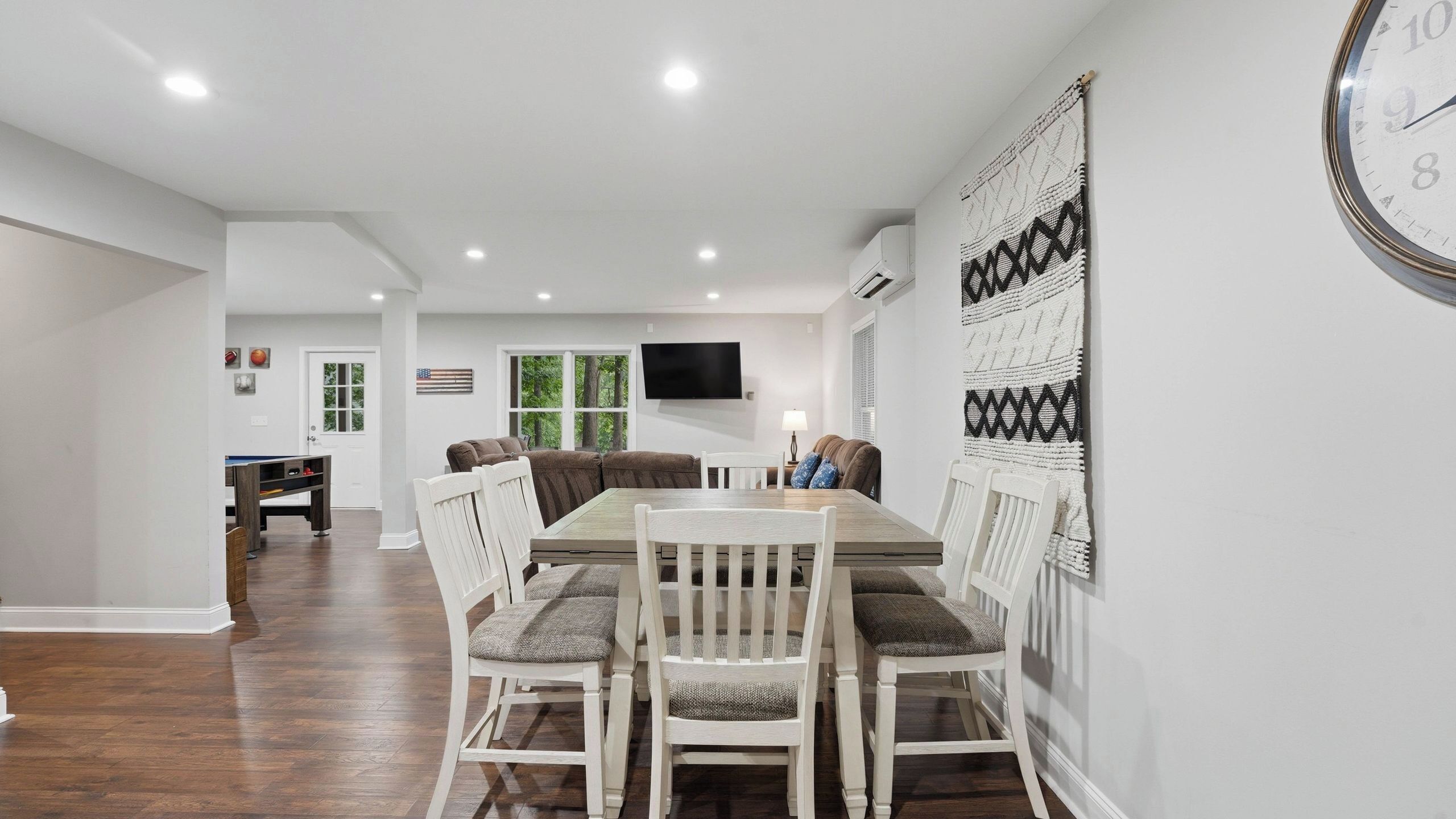 A dining area with a wooden table and chairs, a TV mounted on the wall, and a view of the outdoors through large windows.