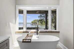 A modern bathroom with a freestanding bathtub, a window with a view of a lake and dock, and a wooden tray with a glass of water. - Water, Water Everywhere by Avantstay Pool + Dock (Mooresville)