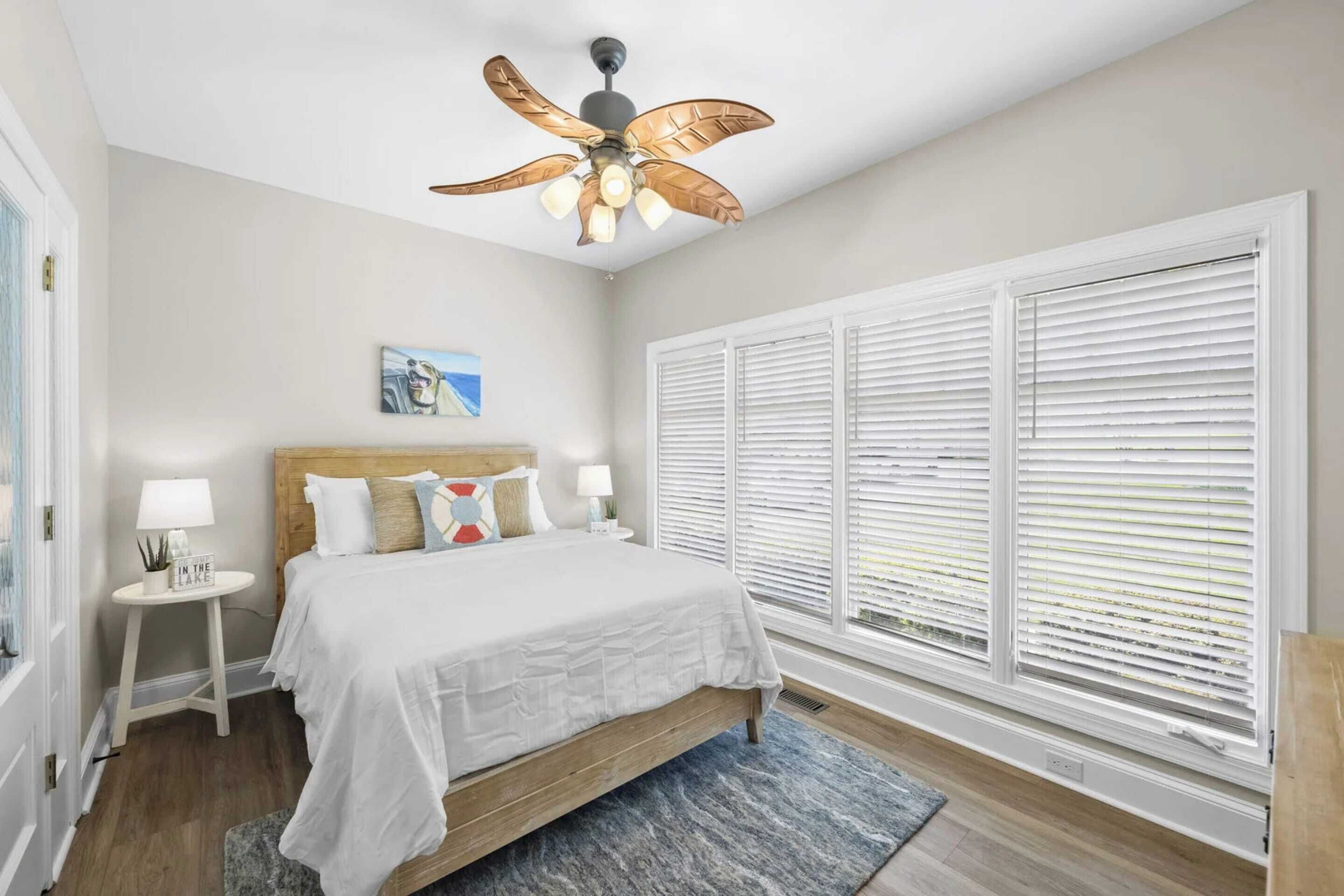A bedroom with a bed, a nightstand, a ceiling fan, and large windows with blinds.