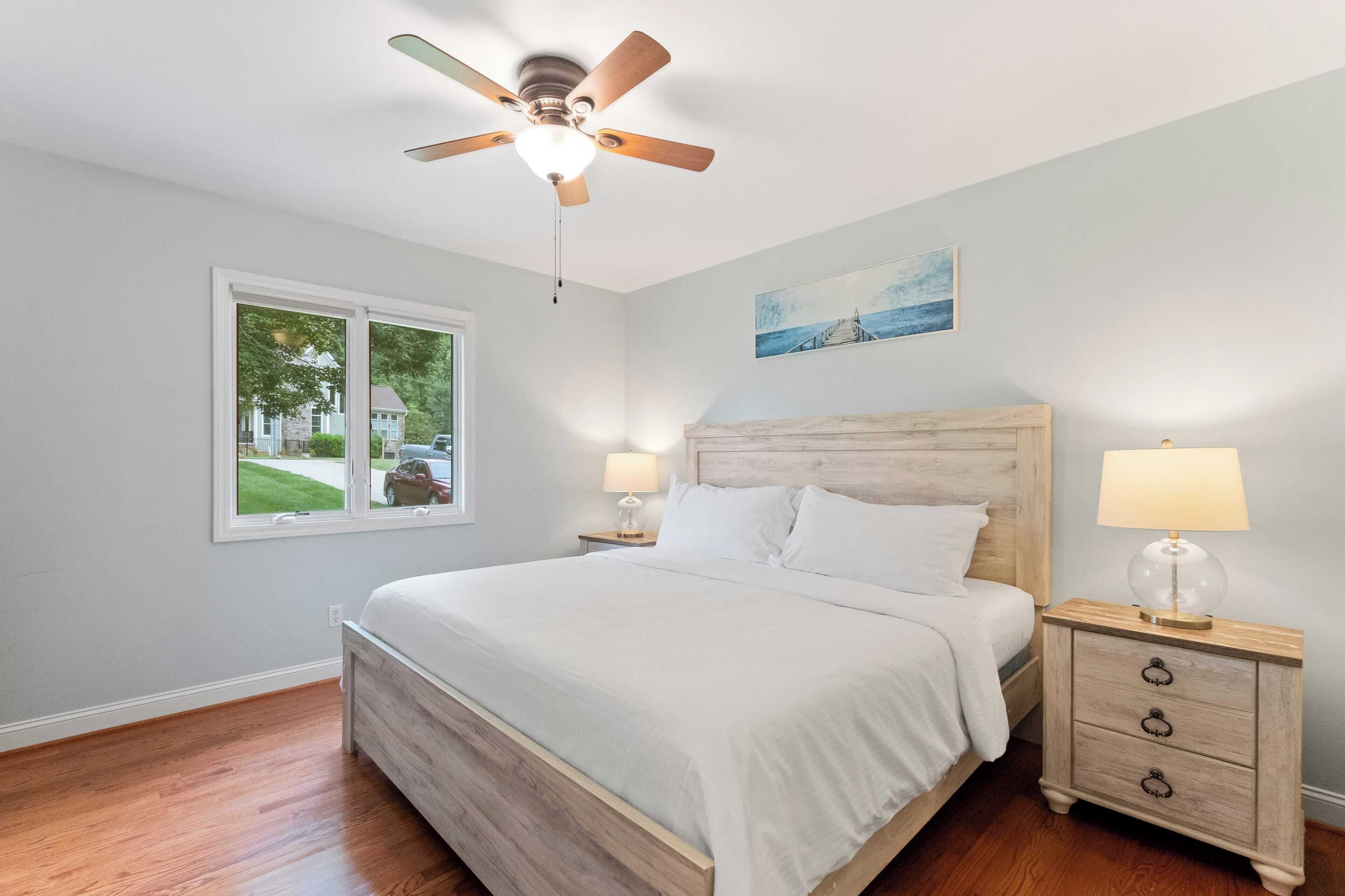 A bedroom with a bed, a nightstand, a ceiling fan, a window, and a painting on the wall.