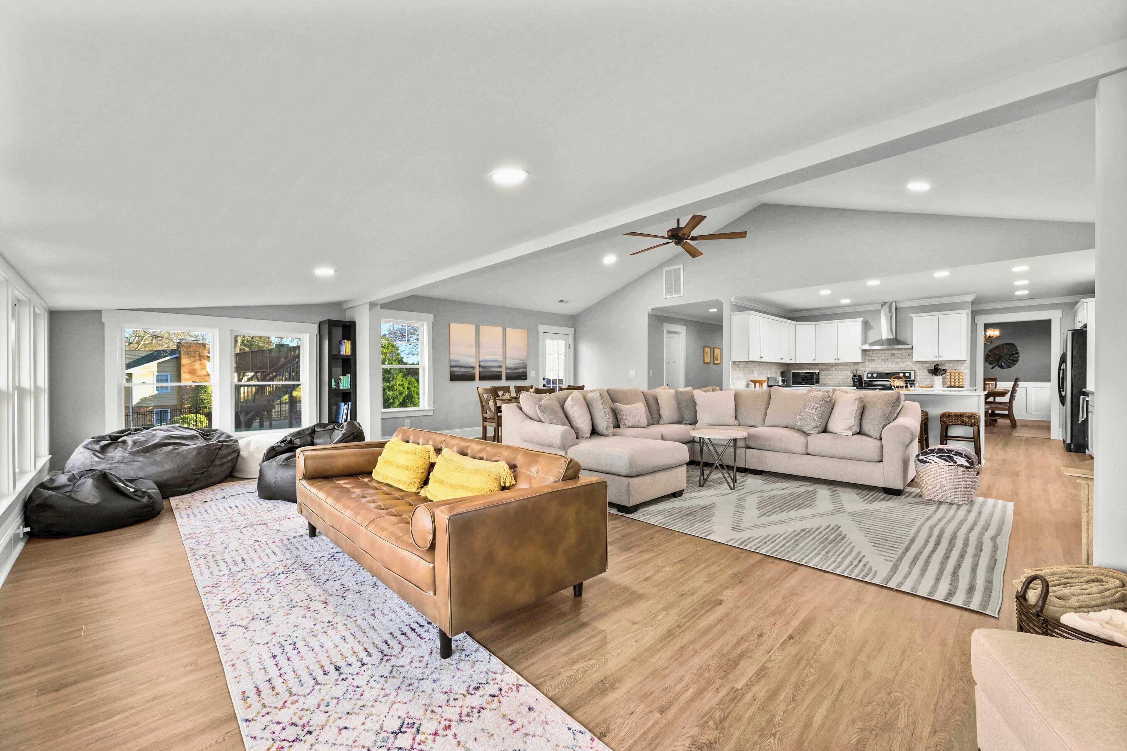 A spacious living room with a large sectional sofa, a coffee table, and a fireplace. There is a kitchen in the background with white cabinets and a ceiling fan.