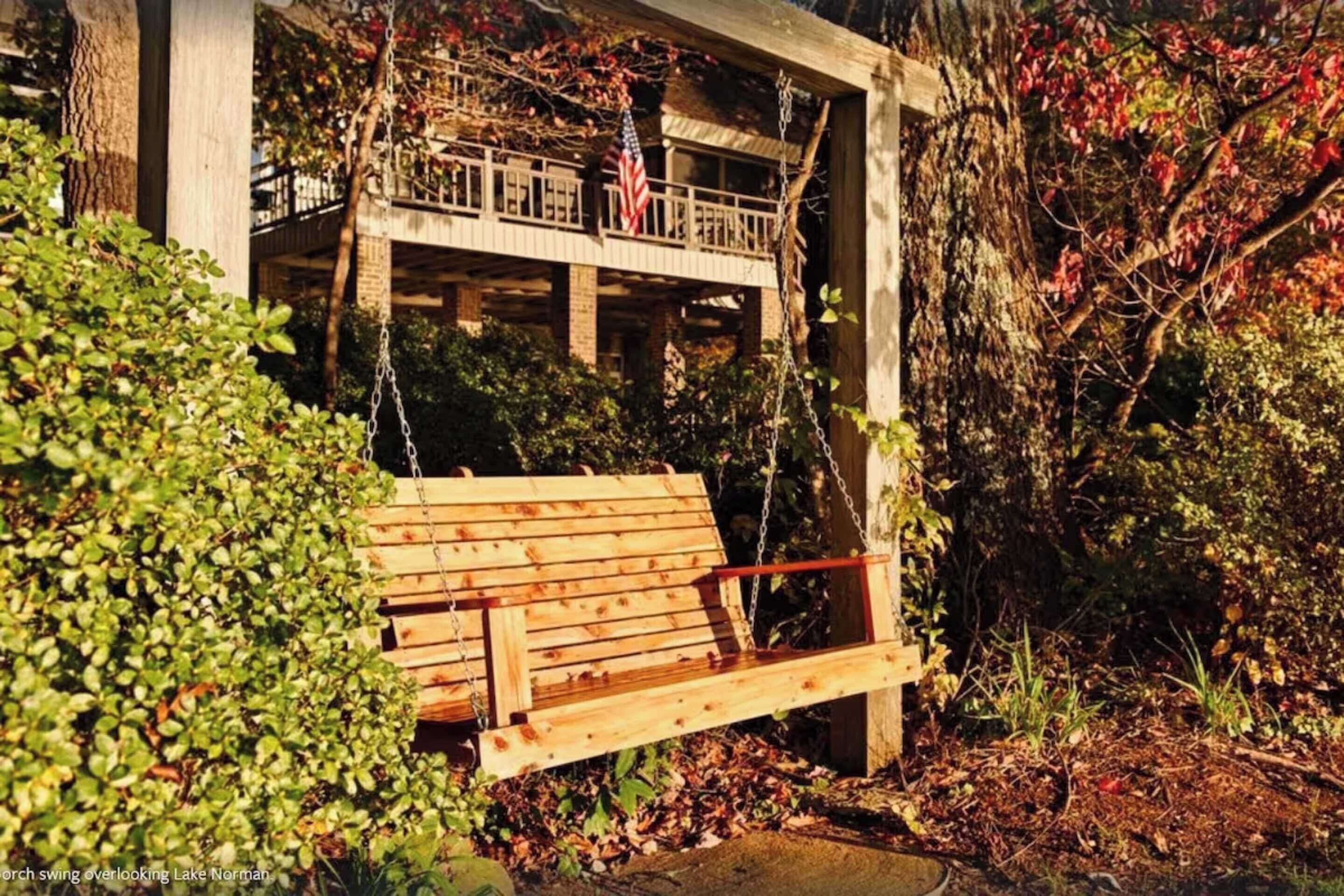Une balançoire en bois surplombe un bâtiment doté d’un balcon, le tout étant entouré d’arbres et de feuillage.