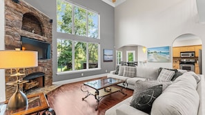 A spacious living room with a stone fireplace, large windows, a sofa, a coffee table, and a kitchen area. - Lakeside Landing by Avantstay Private Dock (Mooresville)