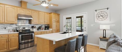 A modern kitchen with wooden cabinets, stainless steel appliances, a central island with bar stools, and a ceiling fan.