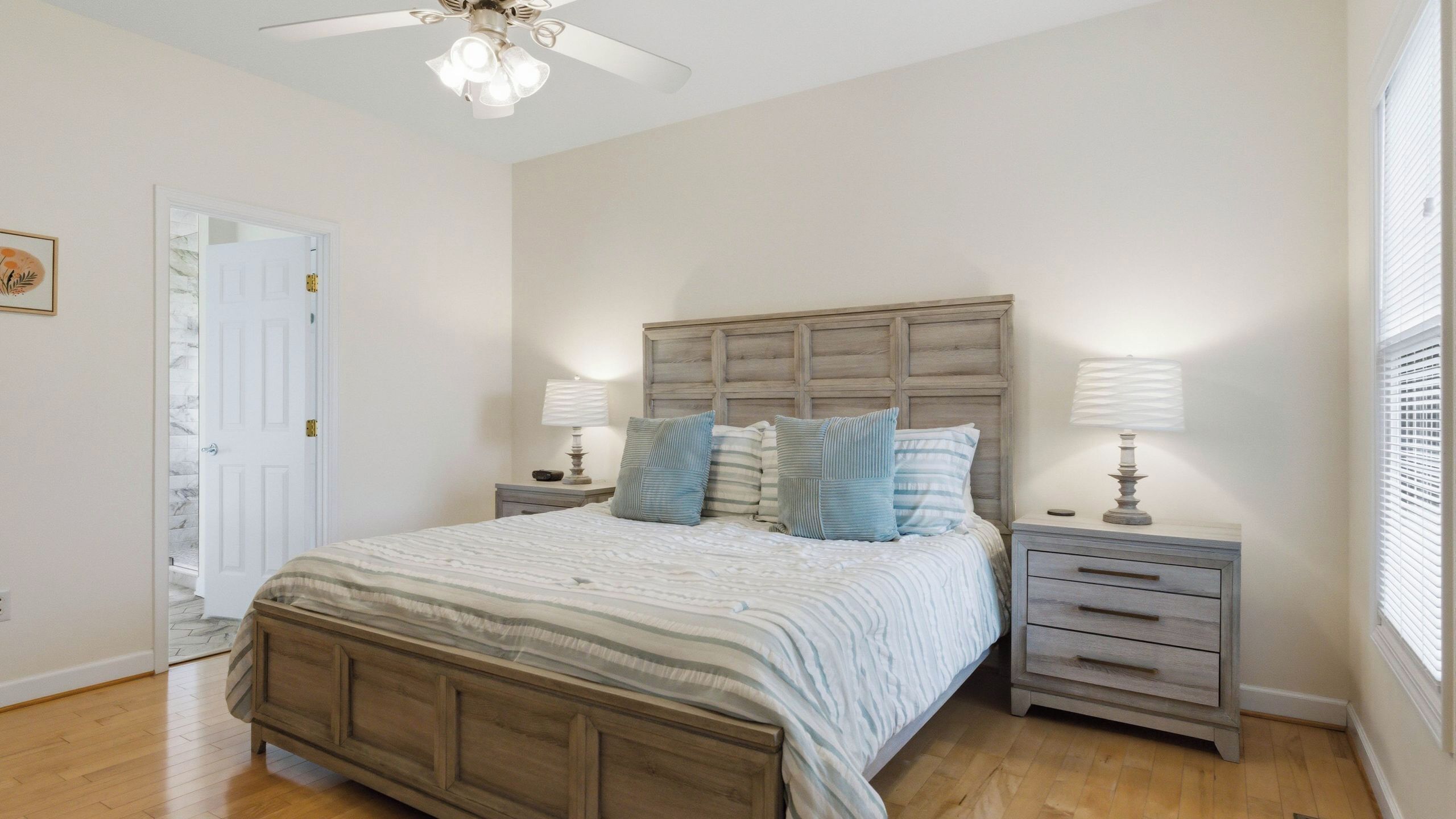 A bedroom with a wooden bed, two bedside tables with lamps, a ceiling fan, and a door leading to another room.