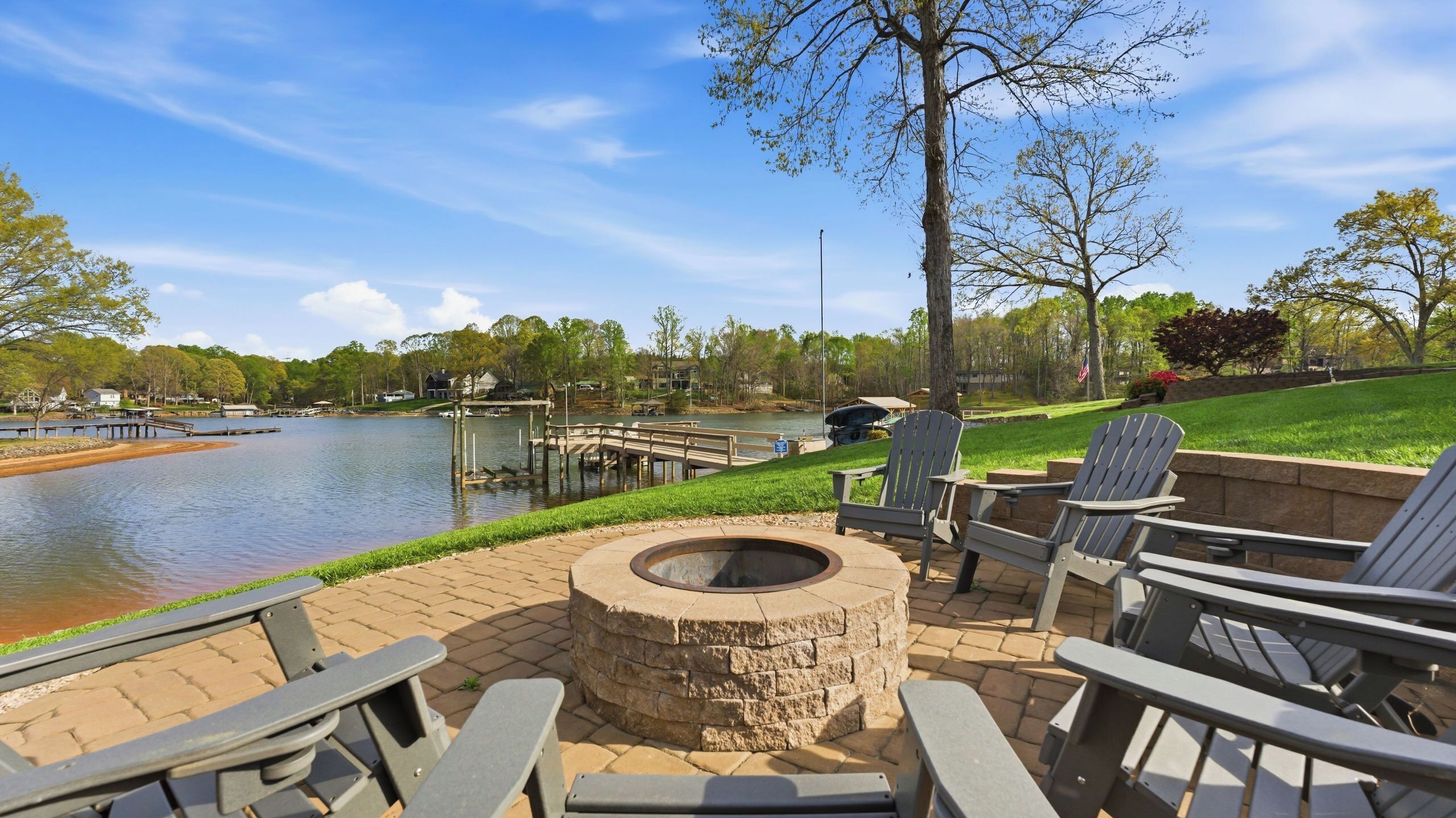 A lakeside patio with Adirondack chairs, a stone fire pit, and a view of the lake.