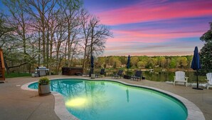 A swimming pool with a curved edge, surrounded by lounge chairs and umbrellas, with trees and a distant view of a house.
