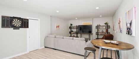 A modern living room with a sofa, a TV, a dartboard, and a dining area with stools.