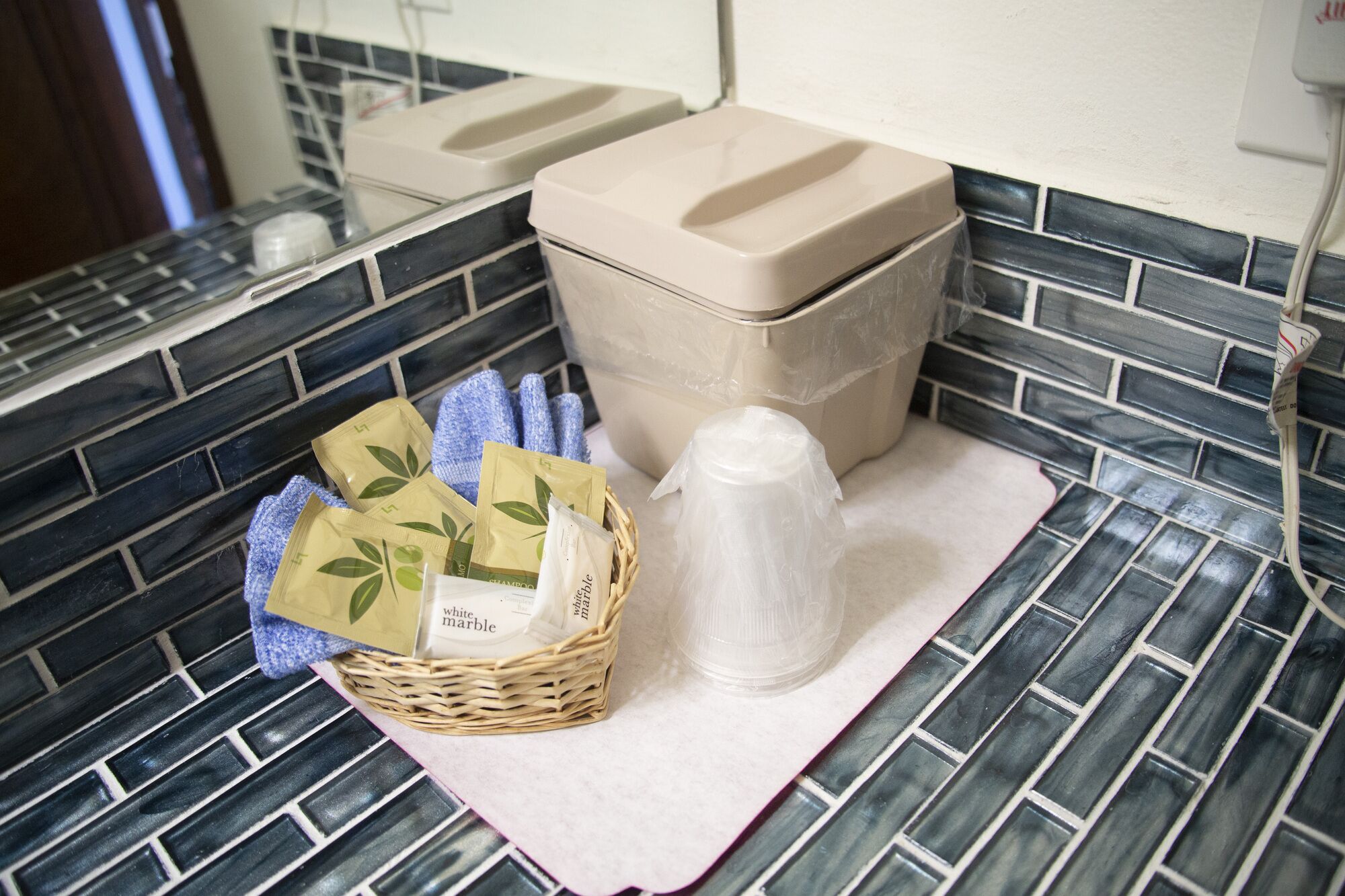 A bathroom with a tiled backsplash, a trash bin, a basket with toiletries, and a plastic bag on a towel.