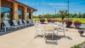En uteplats med solstolar och ett bord, en byggnad i tegel med stora fönster och en välskött trädgård.