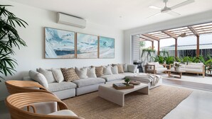 A modern living room with a sectional sofa, a coffee table, and a dining area with a wooden table and chairs. There are two large abstract paintings on the wall, and a ceiling fan is visible.