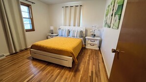Un dormitorio con cama, mesitas de noche, una ventana con cortinas y una puerta.