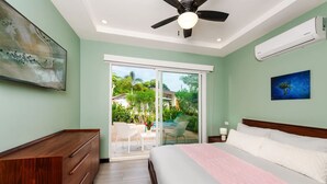 Une chambre avec un lit, un ventilateur de plafond, une table de chevet et une porte-fenêtre coulissante donnant sur un espace extérieur aménagé avec des chaises et une table.